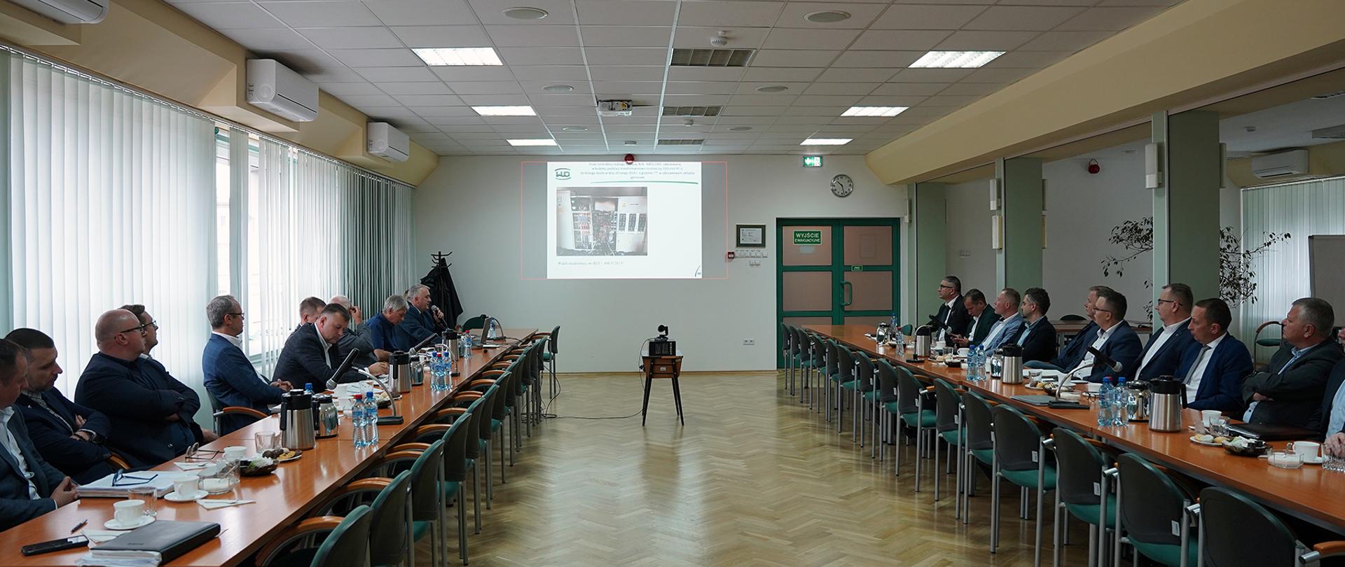 Grupa osób siedzi przy dwóch stołach i spogląda na ekran, na którym wyświetlane są slajdy.