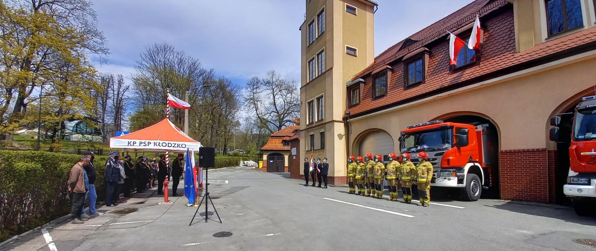 Widok na plac Komendy Powiatowej PSP w Kłodzku. Po lewej stronie uczestniczy uroczystości. Po prawej stronie widok na budynek Komendy Powiatowej PSP w Kłodzku. Przed budynkiem stojący w szeregu strażacy w umundurowaniu specjalnym. 