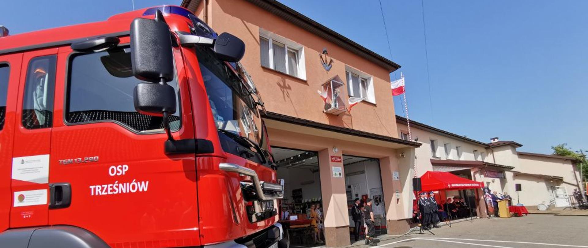 Kolorowa fotografia wykonana w słoneczny dzień przedstawia na pierwszym planie część czerwonego wozu bojowego straży pożarnej a w tle część frontową budynku remizy strażackiej. W otwartych bramach garażowych widać stojących strażaków a w dalszym planie widoczny czerwony namiot – pawilon, pod którym stoją strażacy i osoby cywilne, podczas uroczystej zbiórki.