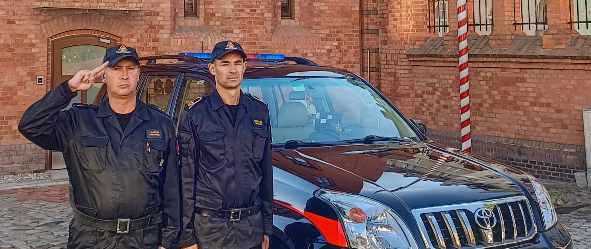 strażacy w czarnych mundurach stoją na baczność, za nimi pojazd pożarniczy, oddanie hołdu