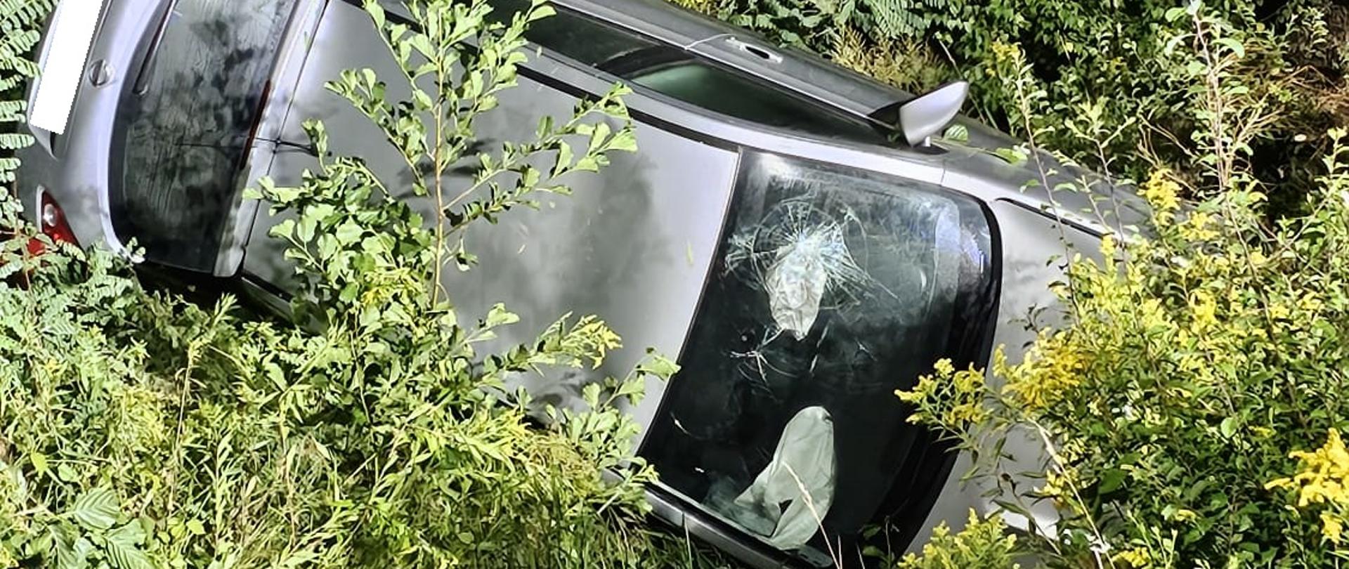 Na zdjęciu widać pojazd osobowy w rowie, pojazd leży na prawym boku, otwarta klapa bagażnika, wokół zarośla i krzaki, poduszki powietrzne zostały wyzwolone, stłuczona przednia szyba na wysokośći kierowcy.