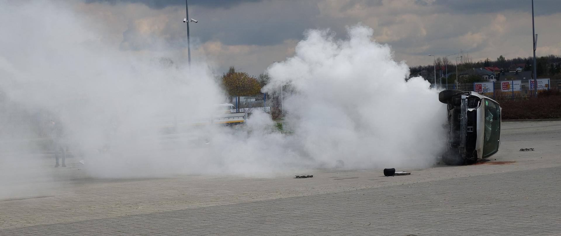 Na zdjęciu przedstawiono dynamiczny pokaz działań służb ratowniczych w trakcie wypadku komunikacyjnego. Samochód osobowy leży przewrócony na bok wśród gęstego dymu imitującym pożar pojazdu.