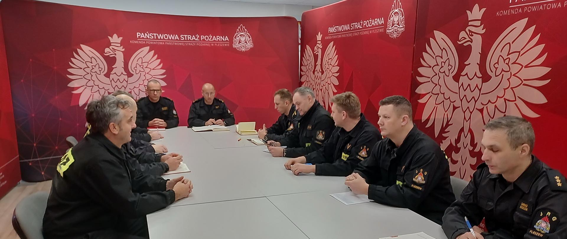 Grupa strażaków siedzi wokół stołu, część z nich ma przed sobą notatniki i długopisy, w tle widoczny baner KP PSP w Pleszewie