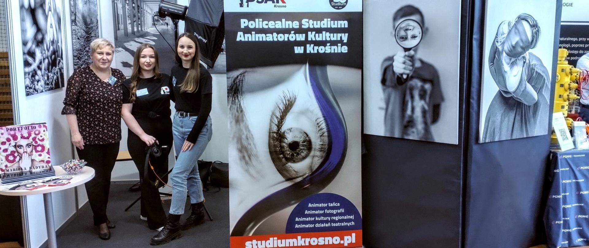 Na zdjęciu stoją dwie słuchaczki Studium wraz z nauczycielką. Stoją na tle czarno-białych zdjęć a po prawej stronie stoi baner reklamowy Studium. 