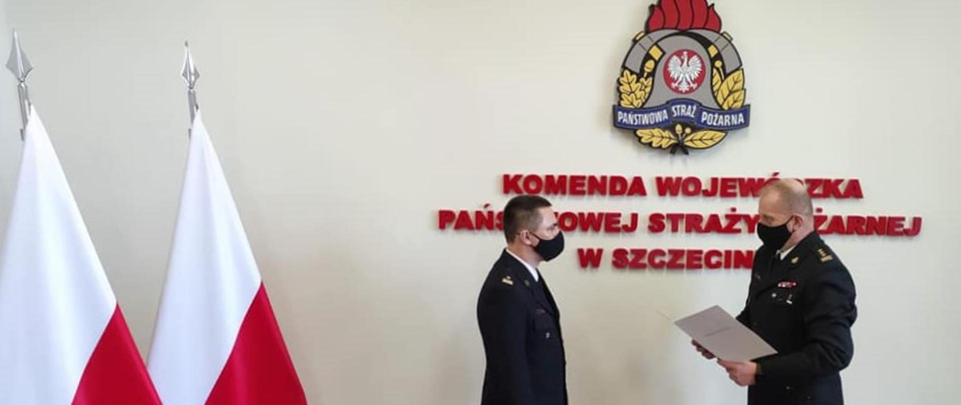 Na zdjęciu dwie osoby ustawione bokiem w mundurach galowych i czarnych maseczkach na twarzy. Po prawej stronie na jasnym tle z umieszczonym na ścianie logotypem Państwowej Straży Pożarnej i czerwonym napisem Komenda Wojewódzka Państwowej Straży Pożarnej stoi Zachodniopomorski Komendant Wojewódzki Państwowej Straży Pożarnej st. bryg. Jarosław Tomczyk odczytując trzymane w dłoni pismo. W centralnej części zdjęcia mł. bryg. Piotr Tuzimek w pozycji wyprostowanej ze wzrokiem na wprost. W lewej części zdjęcia dwie biało-czerwone flagi sztandarowe umieszczone na stojakach flagowych.