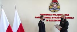 Na zdjęciu dwie osoby ustawione bokiem w mundurach galowych i czarnych maseczkach na twarzy. Po prawej stronie na jasnym tle z umieszczonym na ścianie logotypem Państwowej Straży Pożarnej i czerwonym napisem Komenda Wojewódzka Państwowej Straży Pożarnej stoi Zachodniopomorski Komendant Wojewódzki Państwowej Straży Pożarnej st. bryg. Jarosław Tomczyk odczytując trzymane w dłoni pismo. W centralnej części zdjęcia mł. bryg. Piotr Tuzimek w pozycji wyprostowanej ze wzrokiem na wprost. W lewej części zdjęcia dwie biało-czerwone flagi sztandarowe umieszczone na stojakach flagowych.