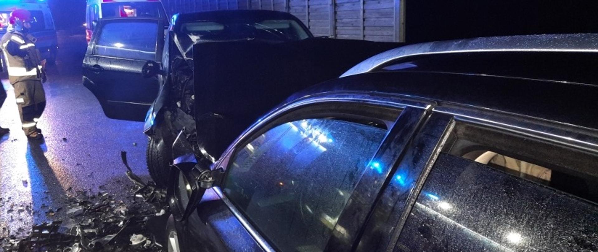 Na zdjęciu widać dwa samochody osobowe które zderzyły się czołowo po lewej stronie stoi strażak w umundurowaniu bojowym w tle dwie karetki pogotowia ratunkowego i ekrany dźwiękochłonne