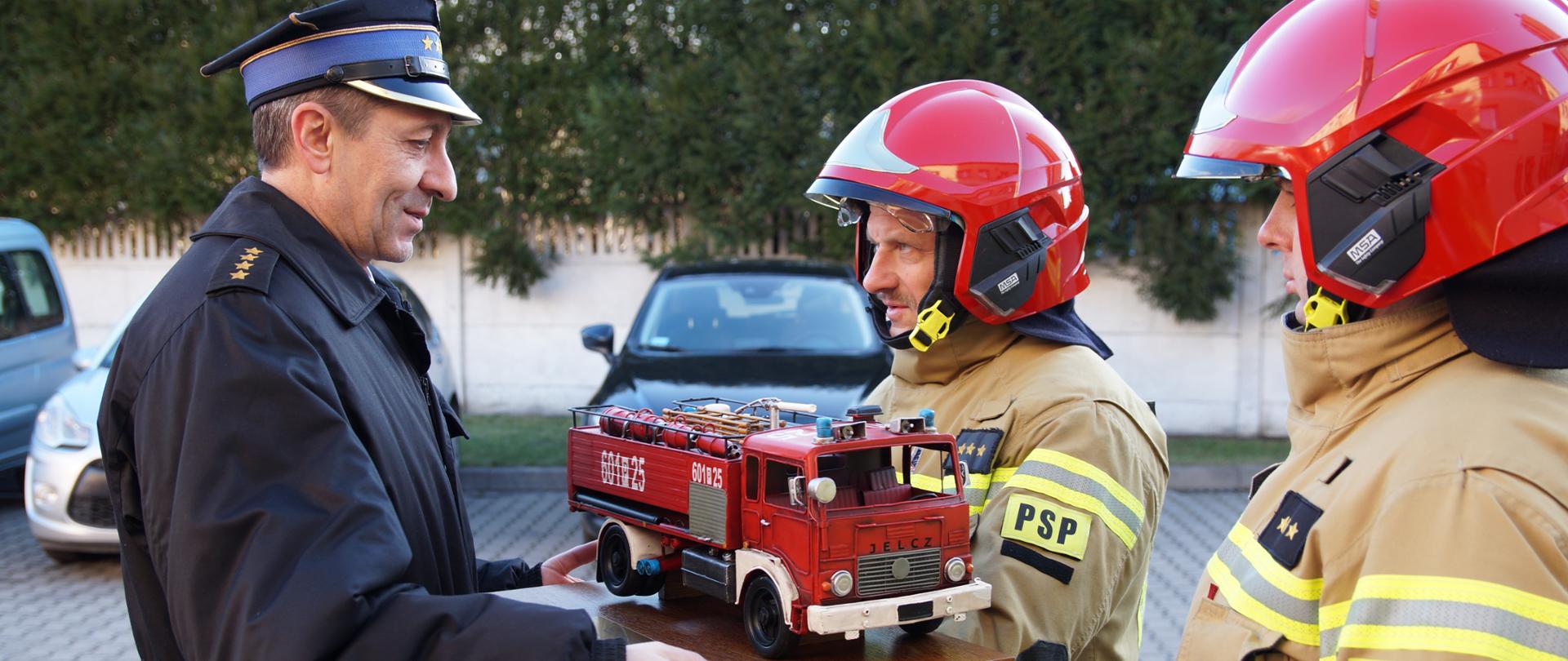 Wręczenie pamiątkowego modelu Jelcza pożarniczego przez jednego z dowódców zmiany służbowej na ręce dowódcy Jednostki Ratowniczo - Gaśniczej odchodzącego na zaopatrzenie emerytalne. 