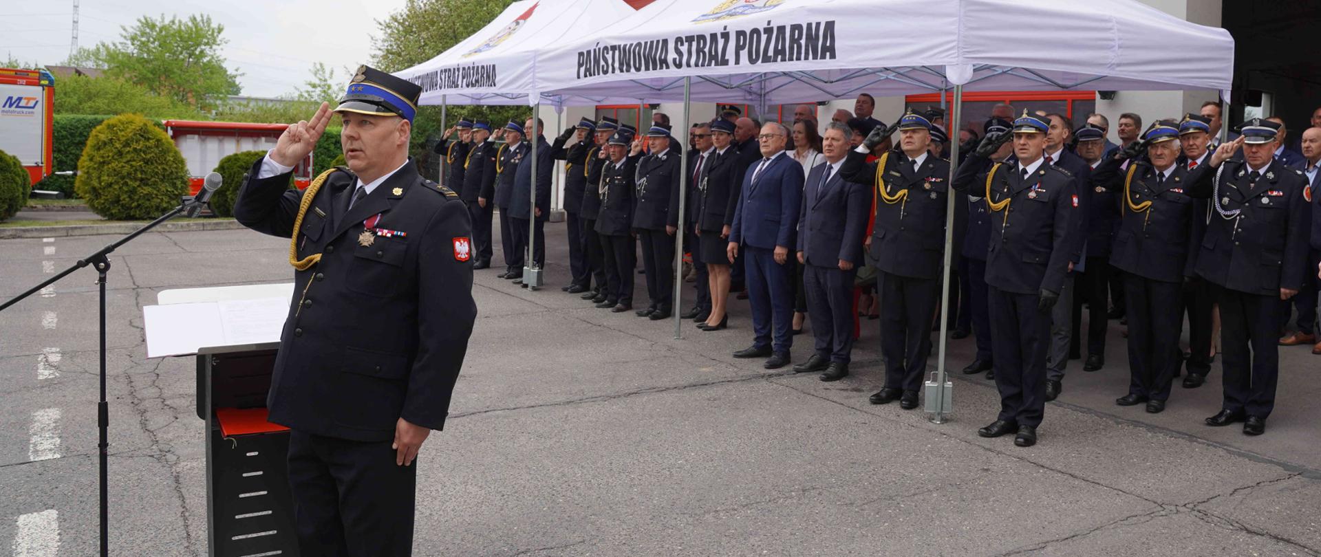Podczas części oficjalnej obchodów Dnia Strażaka, na placu przed budynkiem Państwowej Straży Pożarnej w Opocznie. Prowadzący uroczystość Zastępca Komendanta Powiatowego stoi przy mównicy i oddaje honory. Ubrany jest w galowy mundur strażacki z odznaczeniami. Pod białymi namiotami z napisem „Państwowa Straż Pożarna” stoi komendant powiatowy, komendant wojewódzki oraz zaproszeni goście. Funkcjonariusze ubrani w galowe mundury oddają honory, pozostali stoją na baczność.