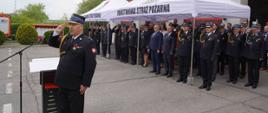 Podczas części oficjalnej obchodów Dnia Strażaka, na placu przed budynkiem Państwowej Straży Pożarnej w Opocznie. Prowadzący uroczystość Zastępca Komendanta Powiatowego stoi przy mównicy i oddaje honory. Ubrany jest w galowy mundur strażacki z odznaczeniami. Pod białymi namiotami z napisem „Państwowa Straż Pożarna” stoi komendant powiatowy, komendant wojewódzki oraz zaproszeni goście. Funkcjonariusze ubrani w galowe mundury oddają honory, pozostali stoją na baczność.