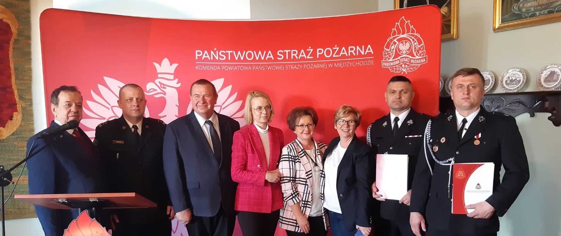 Na zdjęciu widoczna jest uroczystość związana z wręczeniem promes na zakup pojazdów dla OSP. Uroczystość odbywa się wewnątrz pomieszczenia, jest jasno. 