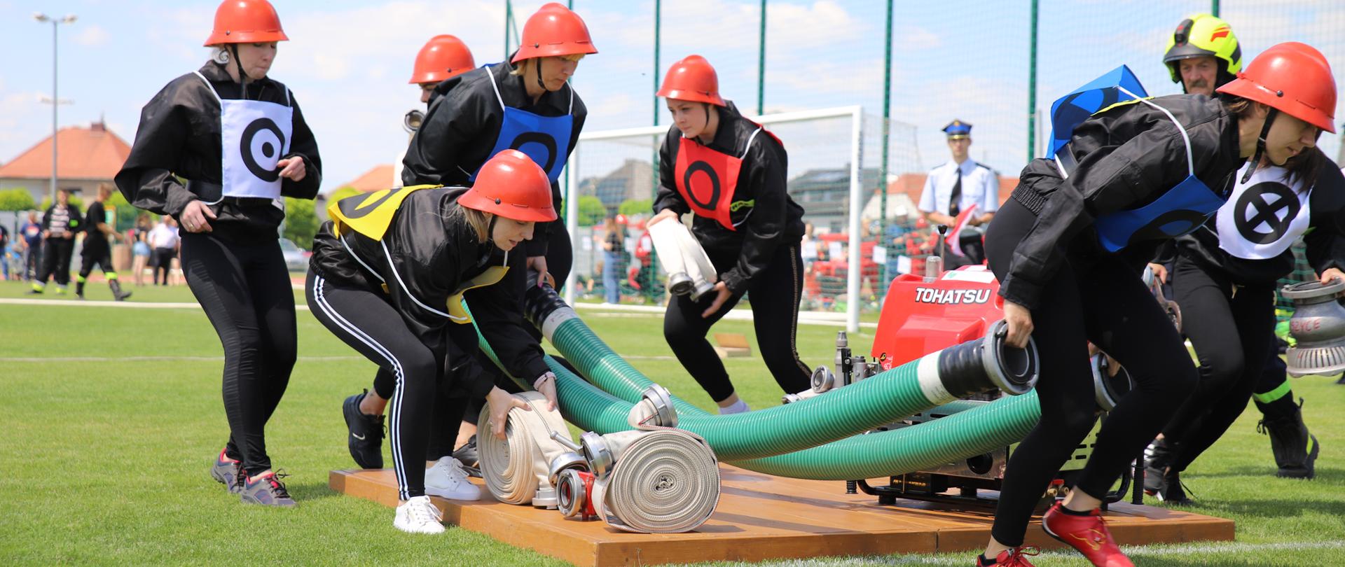 Na zdjęciu dziewczyny w czarnych strojach i czerwonych hełmach zabierają z podestu sprzęt pożarniczy. 