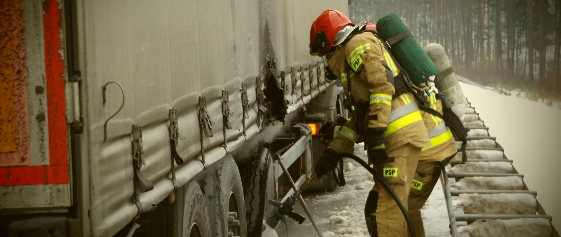 Na zdjęciu znajduje się dwóch strażaków w aparatach ochrony dróg
oddechowych - jeden z nich podaje prąd wody na dymiącą się oponę naczepy.