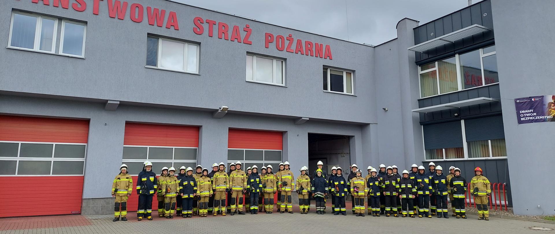 Na zdjęciu widać grupę czterdziestu strażaków, stojących na placu przed Komendą Miejską PSP w Koninie, podczas zbiórki poprzedzającej egzamin szkolenia podstawowego strażaka ratownika Ochotniczych Straży Pożarnych. 
