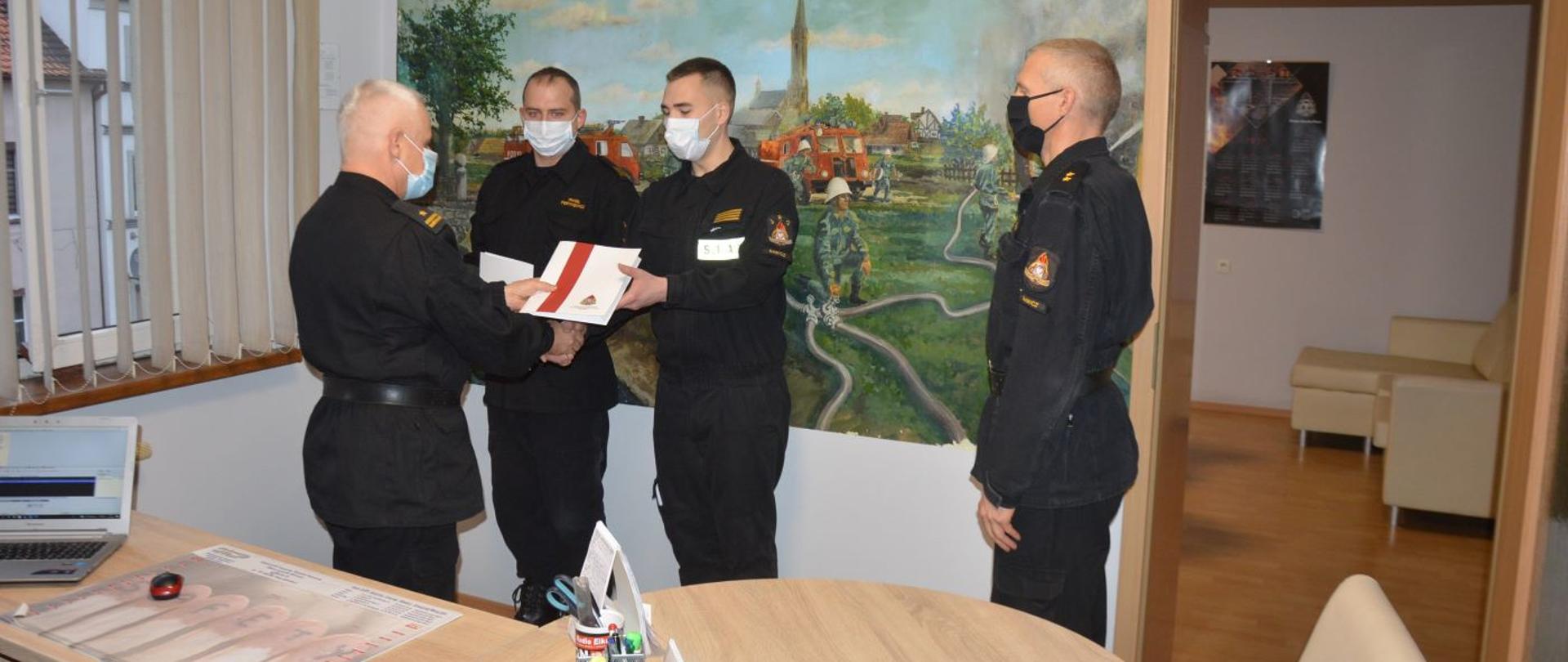 Awanse z okazji Narodowego Święta Niepodległości. Biuro zastępcy komendanta powiatowego PSP w Rawiczu. Zastępca komendanta wręcza akt nadania wyższego stopnia służbowego drugiemu z awansowanych strażaków, składając jednocześnie gratulacje i ściskając jego dłoń. Na tle obrazu ściennego stoją ponadto pierwszy z awansowanych i zastępca dowódcy JRG.