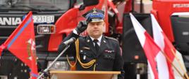 Komendant Powiatowy PSP w Wałczu, st. kpt. Marcin Kulczyk na tle pojazdów pożarniczych oraz flag Polski i Związków Ochotniczych Straży Pożarnej oddaje honor podczas powitania strażaków i zebranych gości