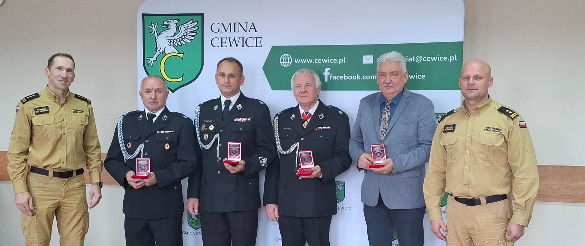 Pamiątkowe zdjęcie wyróżnionych osób pamiątkowym medalem z kadrą kierowniczą Komendy Powiatowej Państwowej Straży Pożarnej w Lęborku. Cztery wyróżnione osoby trzymają w rękach pamiątkowe medale w czerwonym etui. Po lewej i po prawej ich stronie stoi Komendant i Zastępca Komendanta Powiatowego Państwowej Straży Pożarnej w Lęborku. W tle widoczna ścianka baner z napisem Gmina Cewice i herbem gminy.