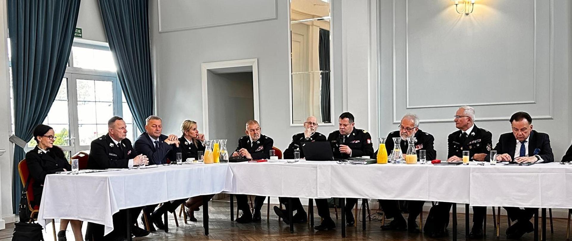 Do mikrofony mówi siedzący za stołem Mazowiecki Komendant Wojewódzki PSP nadbryg. Artur Gonera. Na zdjęciu widocznych kilkunastu innych gości siedzących za stołem. 