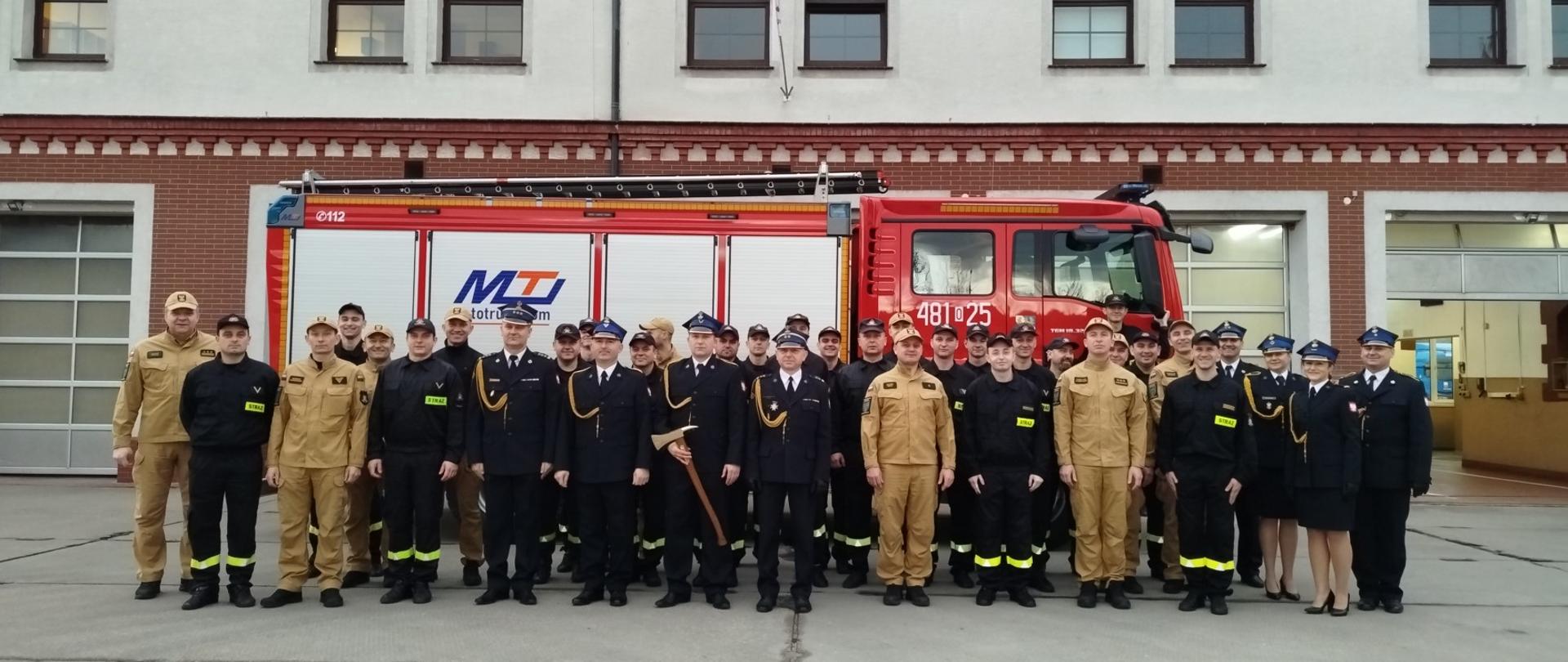 Zdjęcie przedstawia strażaków stojących przed samochodem pożarniczym, w tle budynek strażnicy