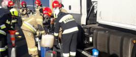 Dwa ciągniki siodłowe wraz z naczepami, stoją w zatoczce autobusowej, strażacy Państwowej Straży Pożarnej oraz Ochotniczych Straży Pożarnych, zbierają wyciekający olej napędowy do wiader, pojemników i baniek, najezdni plama oleju napędowego