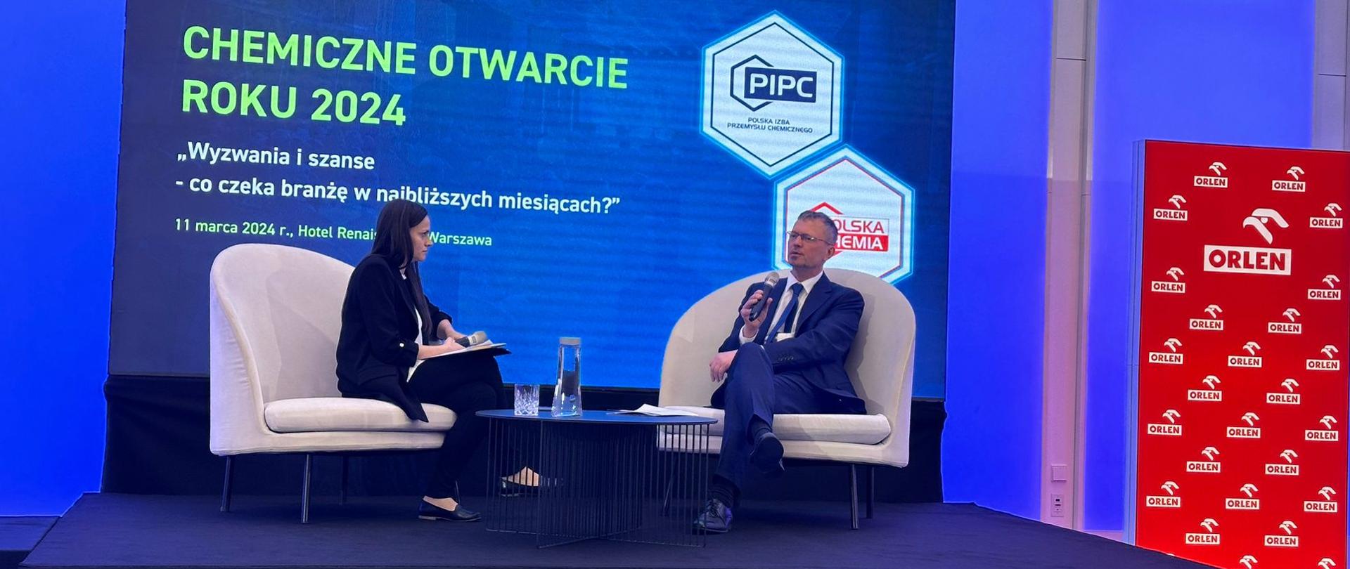 Wiceminister Jacek Bartmiński oraz red. Oksińska siedzą na fotelach, obok stoi stolik. W tle ścianka z napisem Chemiczne Otwarcie Roku 2024 "Wyzwania i szanse - co czeka branżę w najbliższych miesiącach?", 11 marca 2024 r. Po prawej stronie roll up z logotypami Orlenu. 