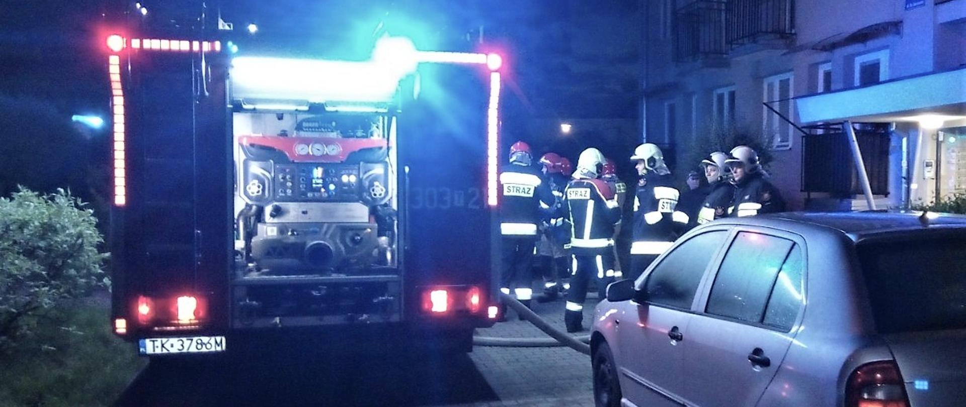 Na zdjęciu przedstawiono miejsce akcji ratowniczo-gaśniczej. Na pierwszym planie, po prawej stronie zaparkowany szary samochód osobowy, po lewej stoi samochód pożarniczy, przy nim grupa strażaków na tle budynku wielorodzinnego, w którym wybuchł pożar.