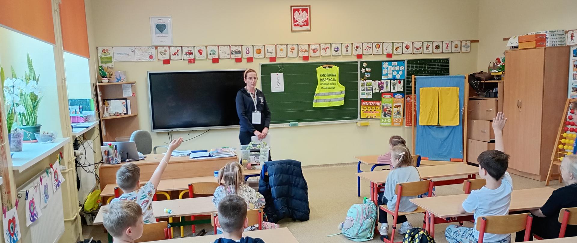Pani Inspektor prowadzi lekcję w klasie, a uczniowie siedzą w ławkach i zgłaszają się, podnosząc ręce; za nią tablica z pomocami dydaktycznymi i zawieszoną żółtą kamizelką z napisem Państwowa Inspekcja Ochrony Roślin i Nasiennictwa 