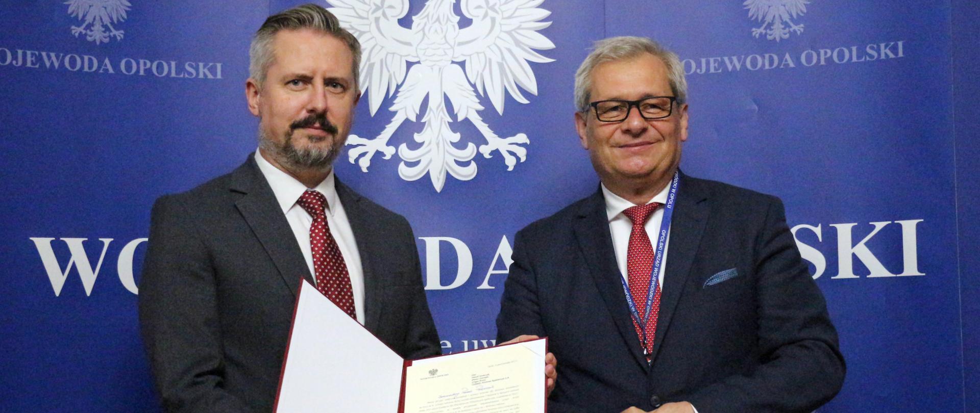 Na zdjęciu Wojewoda opolski i prezes zarządu Grupy Azoty ZAK S.A., pozują do zdjęcia, stojąc obok siebie. Prezes przedsiębiorstwa znajduje się po lewej stronie kadru, trzyma w dłoni teczkę z listem gratulacyjnym od wojewody. Wojewoda stoi po prawej stronie kadru, uśmiecha sie. W tle grantowa ścianka z napisem Wojewoda Opolski oraz orzełkami.