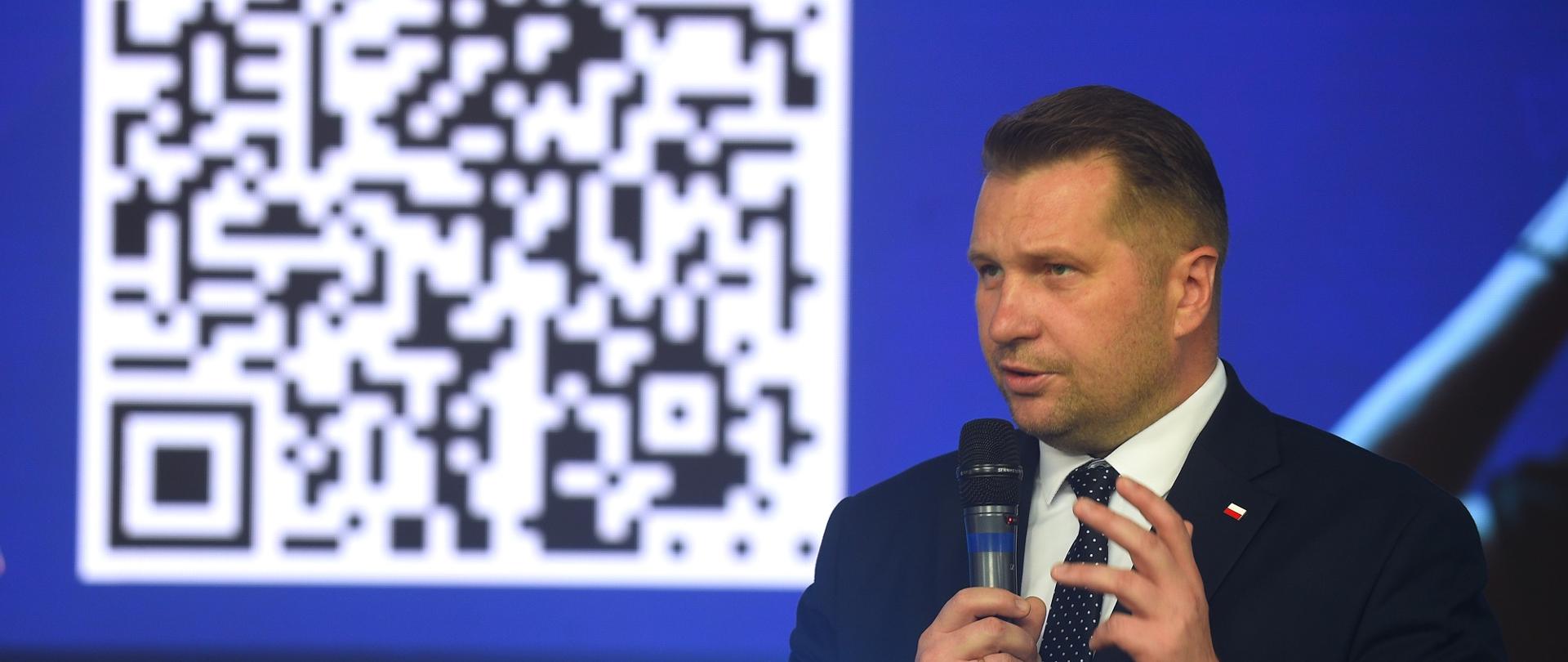 Minister Czarnek mówi do mikrofonu, za nim na wielkim niebieskim ekranie kod QR.