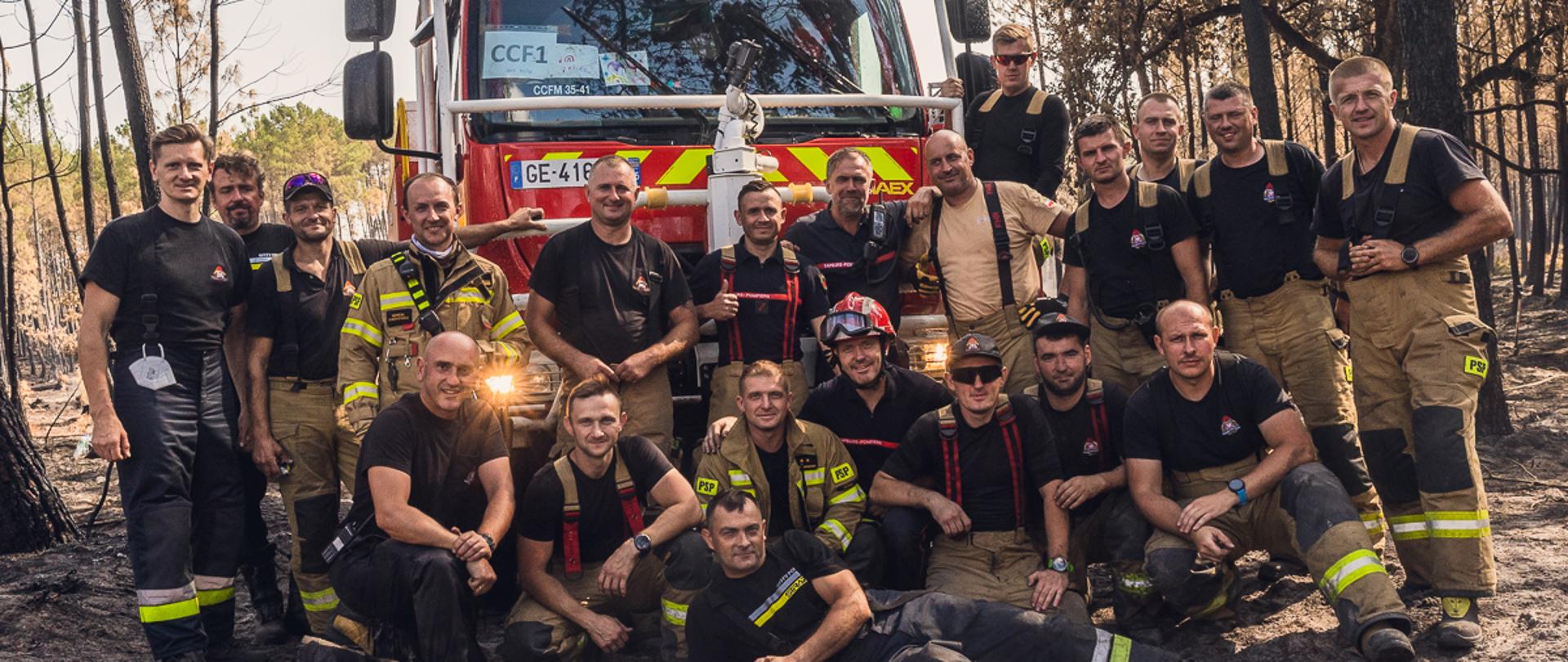 na zdjęciu widać grupę strażaków w ubraniach bojowych, na tle francuskiego samochodu strażackiego. Polscy strażacy są w działaniach gaśniczych w lasach w okolicy Bordeaux (Francja)