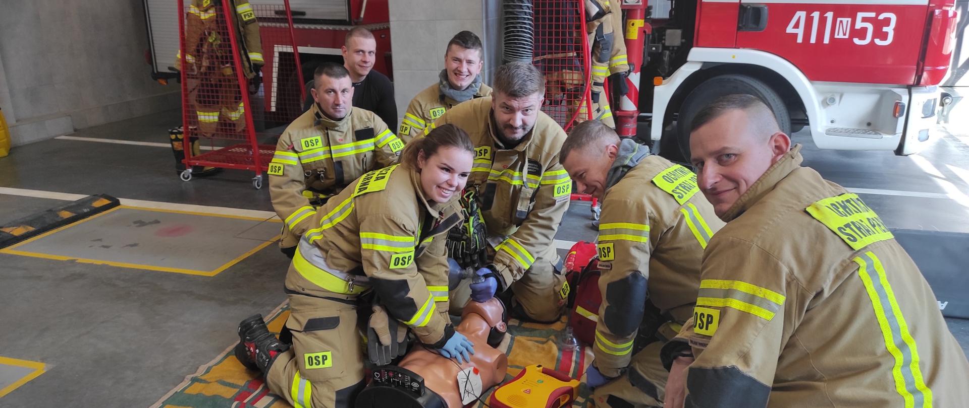 Grupa sześciu strażaków ćwiczy przy manekinie leżącym na podłodze garażu bojowego.