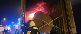 Zdjęcie przedstawia ogień rozprzestrzeniający się w kontenerze oraz strażaków przystępujących do akcji gaśniczej