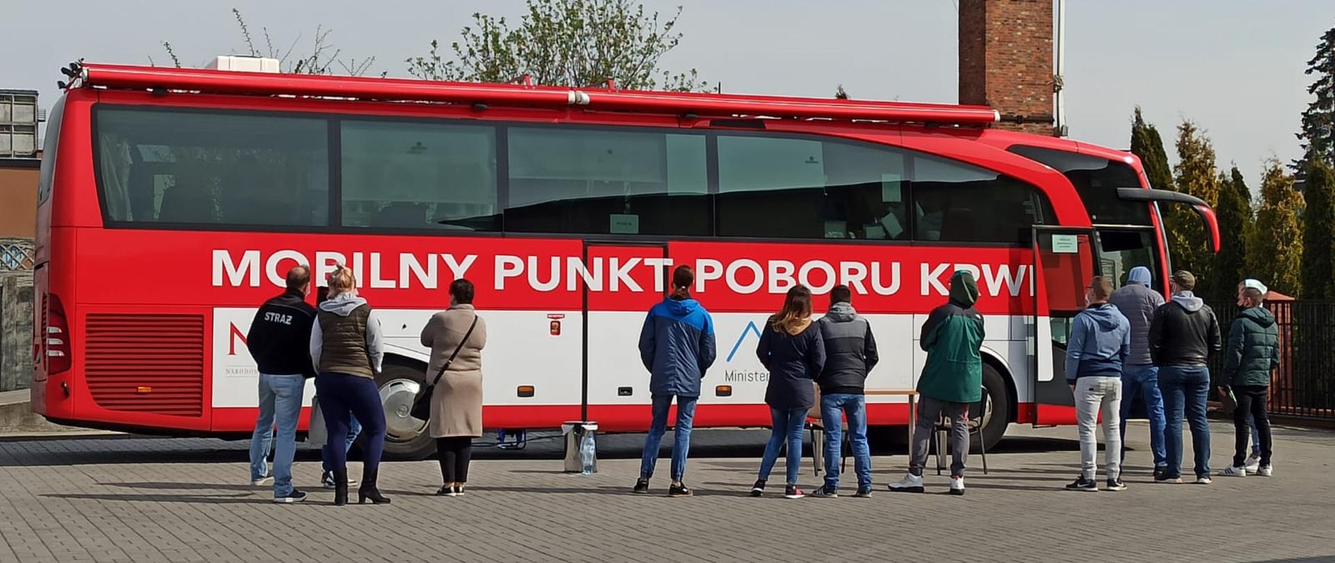 Zdjęcie przedstawia autobus krwiodawstwa oraz zgromadzonych przed nim ludzi. Autobus jest koloru czerwonego i znajduje się na nim biały napis: Mobilny punkt poboru krwi. Osoby zgromadzone przed autobusem stoją w kolejce do oddania krwi, wszyscy ustawieni są plecami do zdjęcia. Za autobusem, w tle zdjęcia widać ceglany komin oraz rosnące drzewa i tuje. 