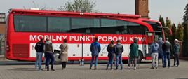 Zdjęcie przedstawia autobus krwiodawstwa oraz zgromadzonych przed nim ludzi. Autobus jest koloru czerwonego i znajduje się na nim biały napis: Mobilny punkt poboru krwi. Osoby zgromadzone przed autobusem stoją w kolejce do oddania krwi, wszyscy ustawieni są plecami do zdjęcia. Za autobusem, w tle zdjęcia widać ceglany komin oraz rosnące drzewa i tuje. 
