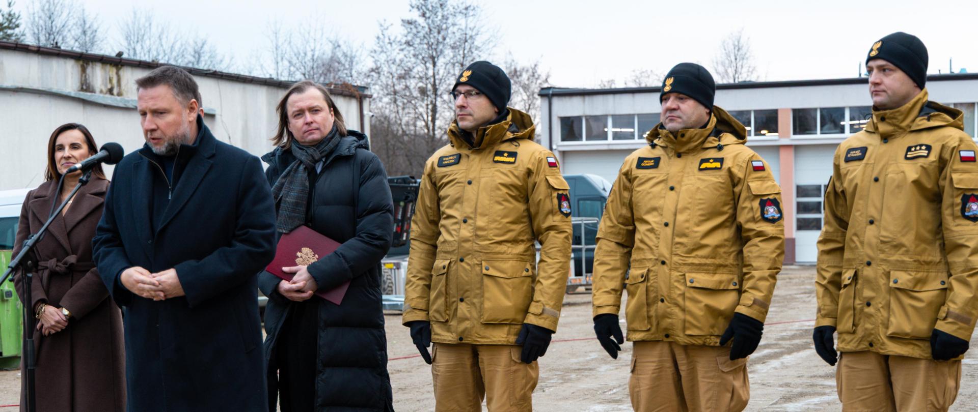 Mężczyzna przemawia do mikrofonu na zewnątrz jednostki PSP. Obok niego stoi kobieta i mężczyzna z dokumentem, a po drugiej stronie trzech strażaków w żółtych mundurach PSP.