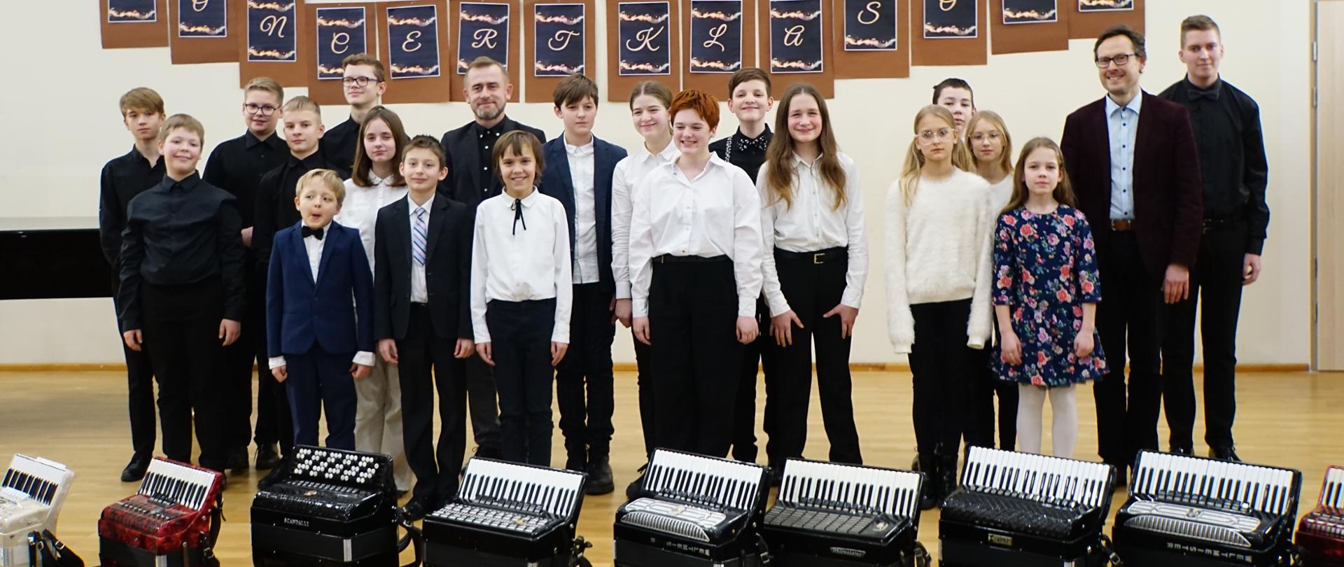 Na sali koncertowej stoją ustawieni wszyscy uczniowie obu klas akordeonu wraz z nauczycielami. Pozują do zdjęcia. Przed nimi na podłodze stoją ustawione w rzędzie akordeony. Powyżej napis "Państwowa Szkołą Muzyczna I st. im. Fryderyka Chopina w Wejherowie" oraz "Koncert klasowy".