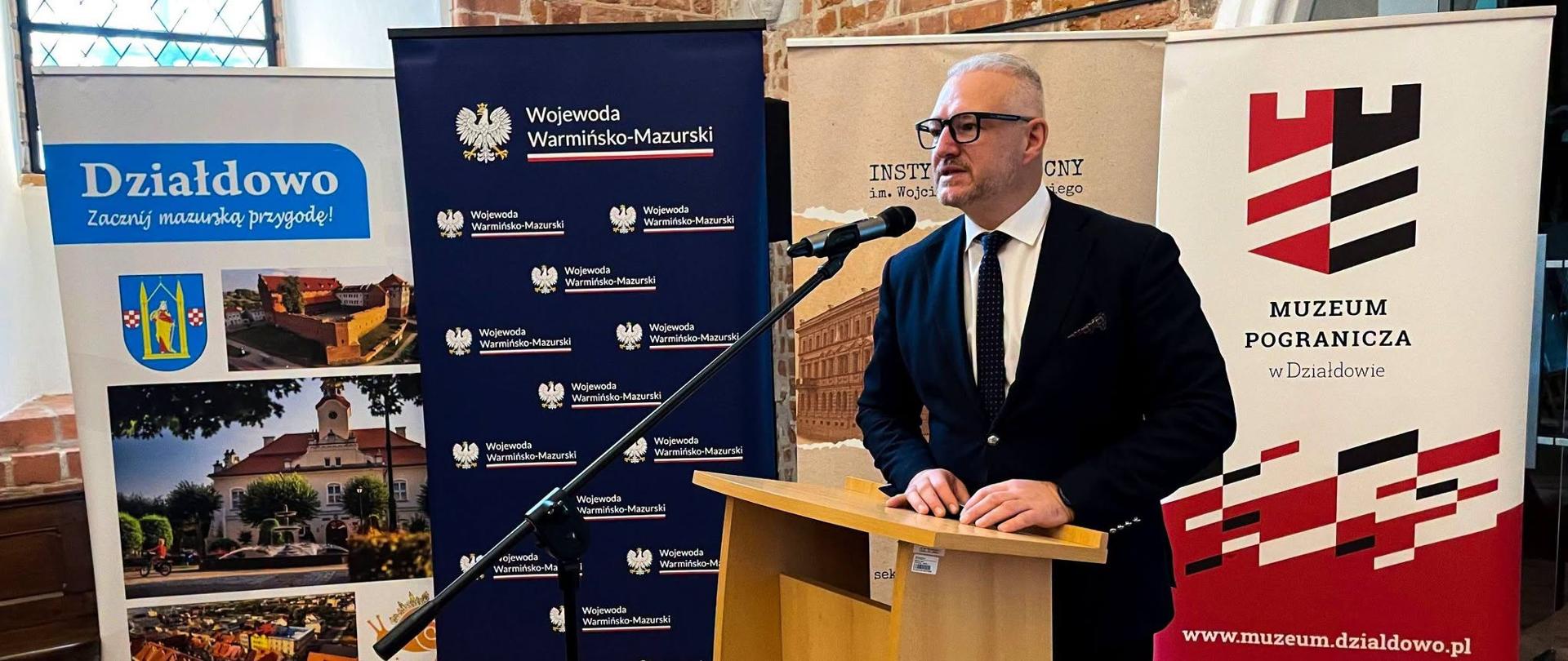 Mężczyzna w granatowym garniturze i okularach, wojewoda, przemawia przy mównicy na tle ścianki "Wojewoda Warmińsko-Mazurski"