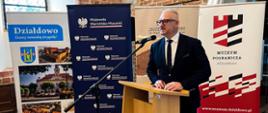 Mężczyzna w granatowym garniturze i okularach, wojewoda, przemawia przy mównicy na tle ścianki "Wojewoda Warmińsko-Mazurski"