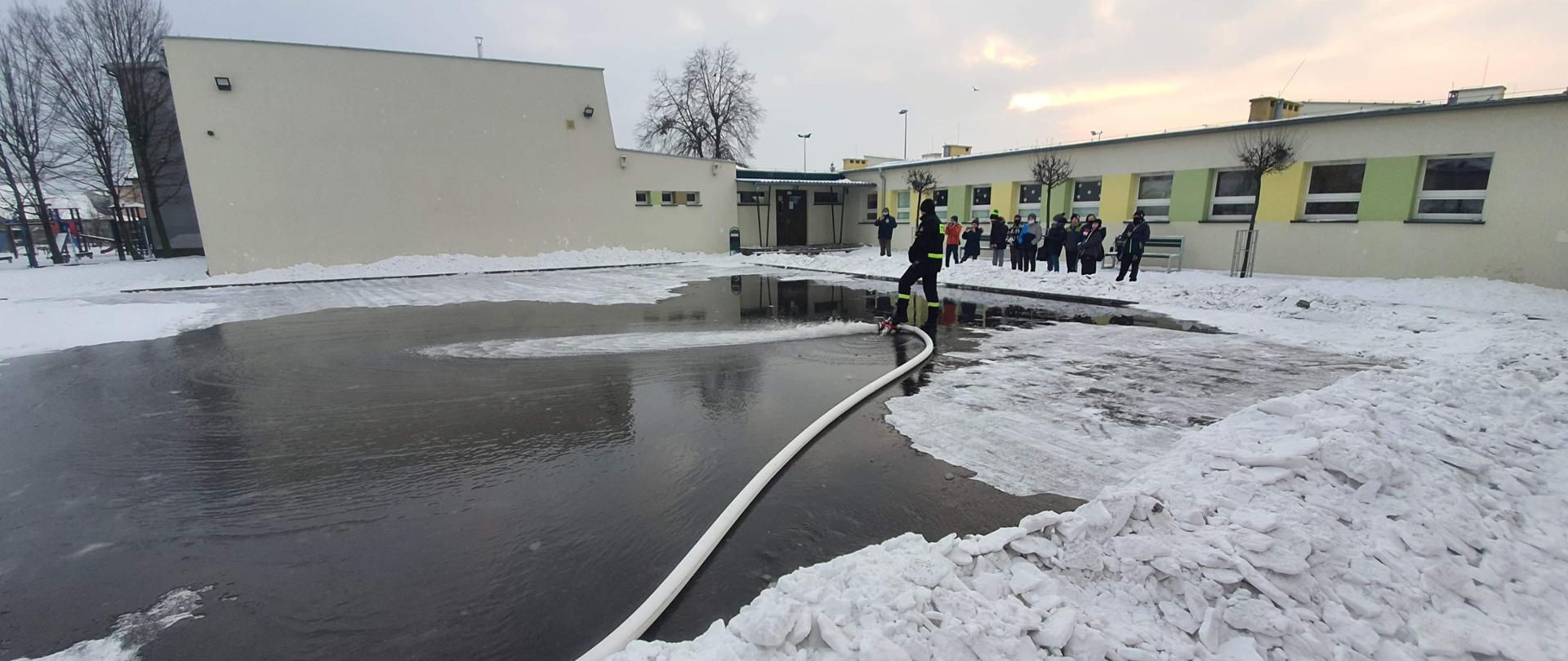 Strażacy wykonują zrzut wody na przygotowanym placu celem utworzenia bezpiecznego lodowiska. Lodowisko znajduje się na terenie szkoły podstawowej. Dzieci przyglądają się strażackiej akcji.