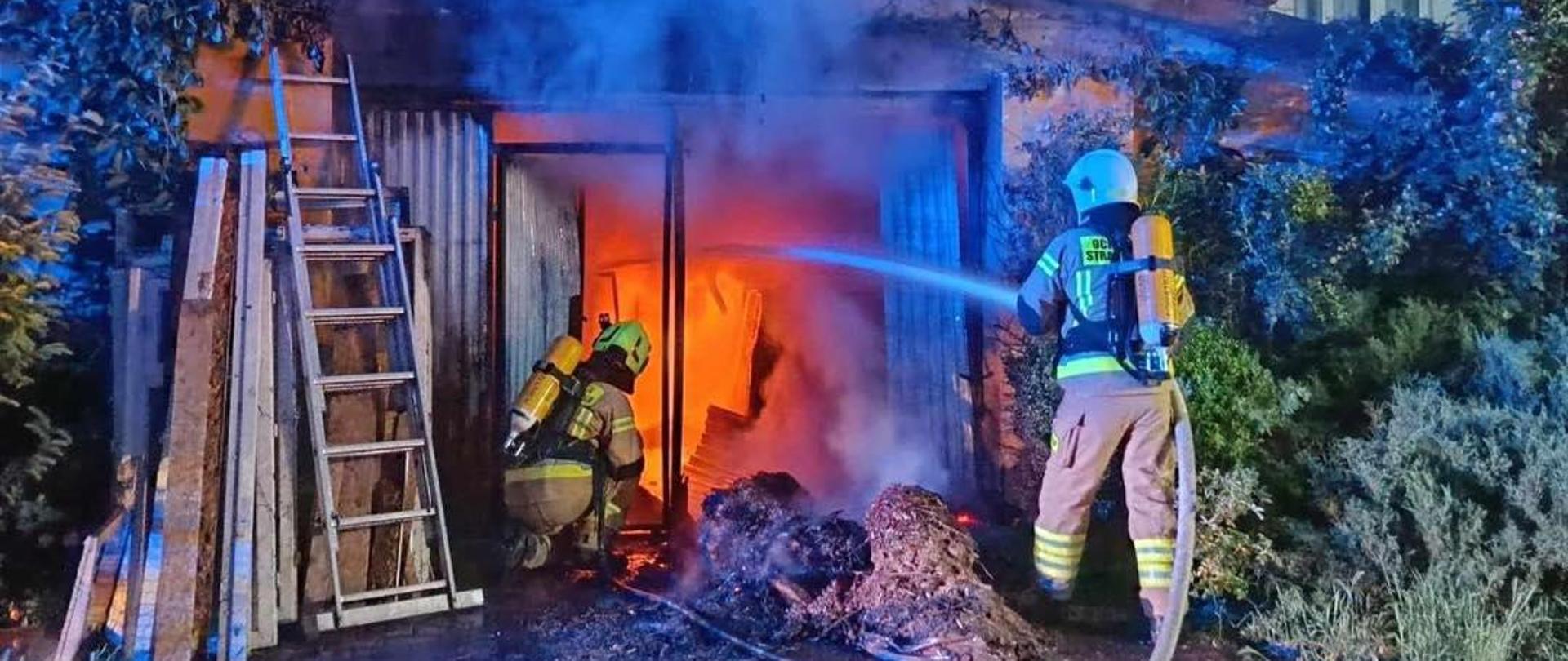 Na zdjęciu widocznych jest dwóch strażaków wyposażonych w aparaty ochrony układu oddechowego. Strażacy podają prądy wody do wnętrza budynku gospodarczego. Wewnątrz budynku gospodarczego widoczny jest pożar. 