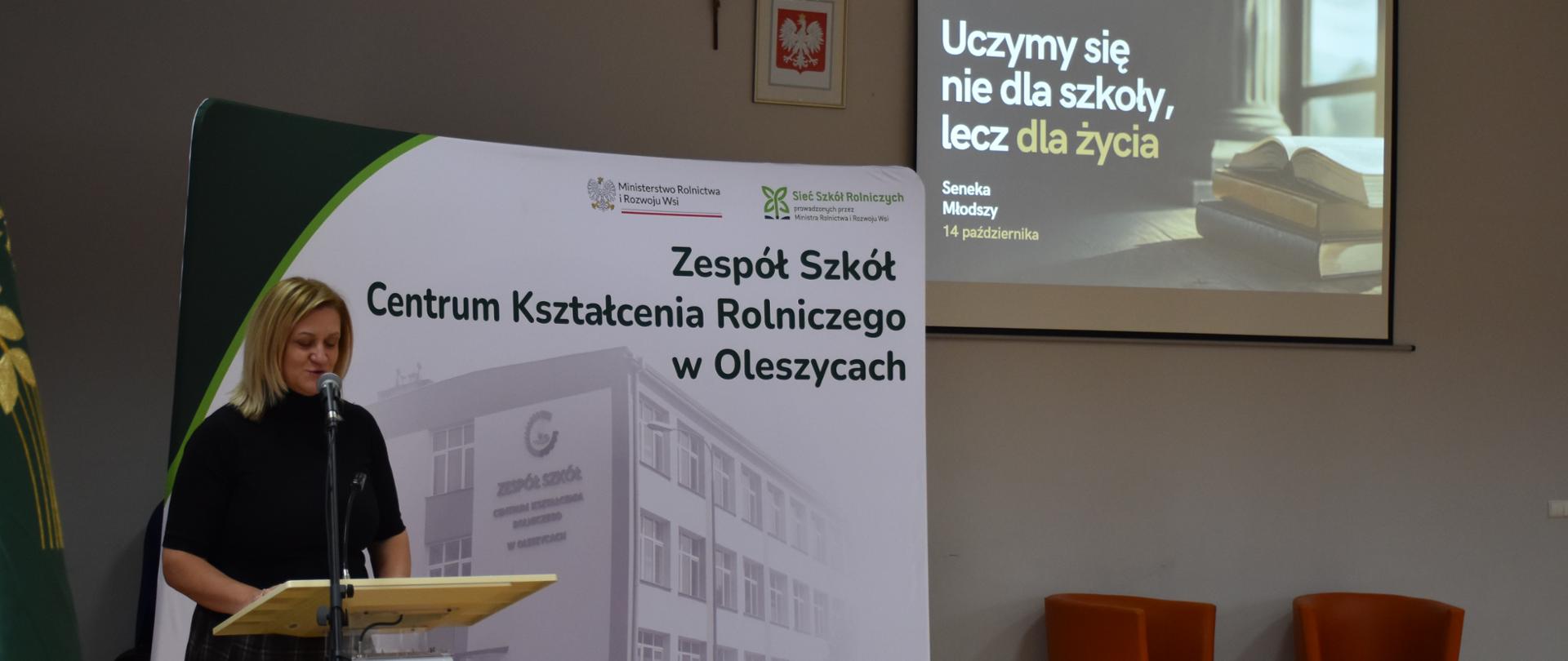 Zdjęcie przedstawia oficjalną uroczystość w Zespole Szkół Centrum Kształcenia Rolniczego w Oleszycach, podczas której kobieta przemawia przy mównicy na tle pamiątkowego baneru. W tle widoczny jest ekran z cytatem Seneki na temat nauki dla życia, a na pierwszym planie znajduje się fragment szkolnego sztandaru.
