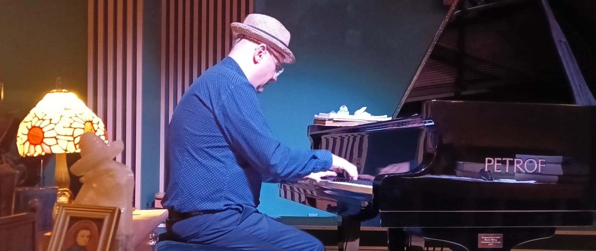 Na zdjęciu znajduje się mężczyzna grający na fortepianie marki PETROF. Jest ubrany w
ciemnoniebieską koszulę, granatowe spodnie i czarną elegancką parę butów. Na głowie ma
jasnobrązowy kapelusz. Siedzi na czarnym stołku i koncentruje się na grze.
Otoczenie sugeruje kameralne wnętrze – widoczne są elementy dekoracyjne, takie jak
stylowa lampa z abażurem witrażowym, rośliny doniczkowe i stolik z obrazkiem oraz figurką.
Ściany mają eleganckie pionowe panele. Oświetlenie tworzy przytulny nastrój, co wskazuje
na miejsce przystosowane do koncertów lub muzycznych spotkań.
