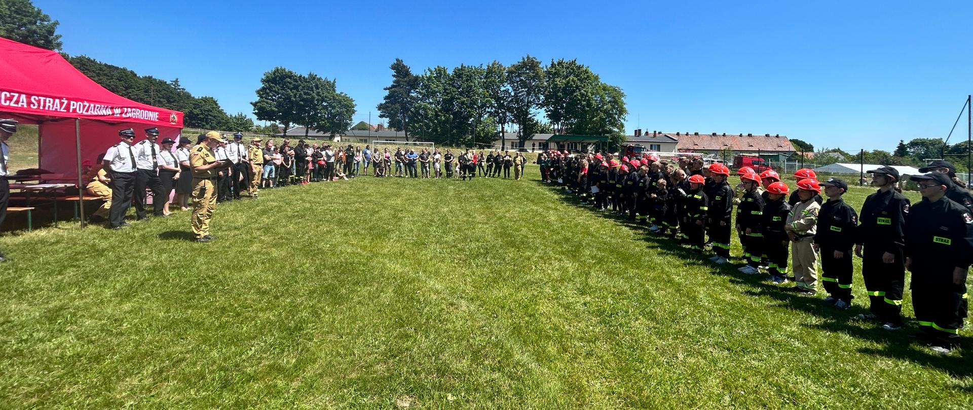 młodzieżowe drużyny pożarnicze na murawie boiska na zbiórce podczas odczytania protokołu z zawodów oraz wręczenia nagród, bezchmurne niebo, w tle drzewa