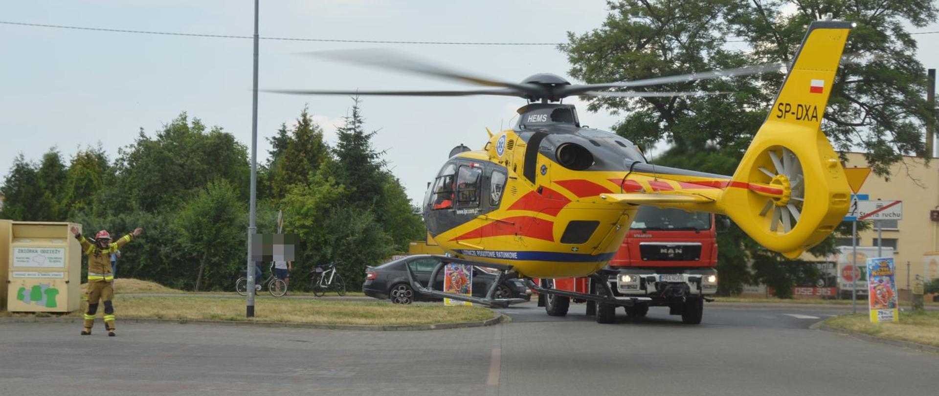 Wspólna akcja strażaków i LPR. Parking jednego z rawickich hipermarketów. Na nim strażak wydający sygnały gestowe pilotowi śmigłowca Lotniczego Pogotowia Ratunkowego, który podchodzi do lądowania. Przed śmigłowcem stoi wóz straży pożarnej blokujący wjazd na parking.