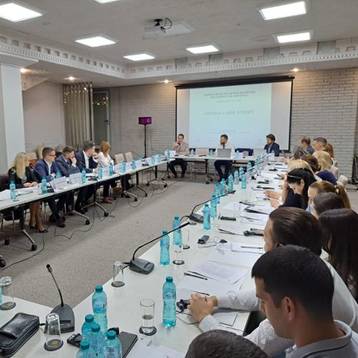 Uczestnicy warsztatów słuchają jednego z trzech prowadzących siedzących u szczytu stołu, za którym ustawiony jest ekran z wyświetlaną prezentacją