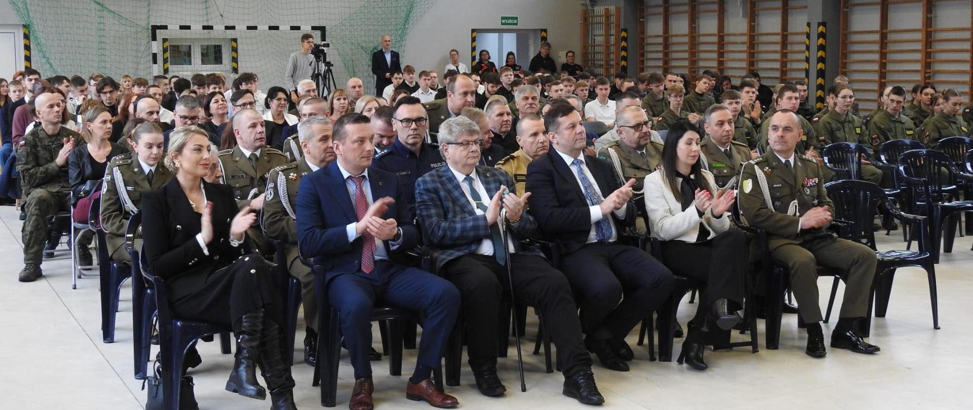 Sala gimnastyczna. Na sali na krzesełkach siedzą przedstawiciele samorządu oraz przedstawiciele służb mundurowych.