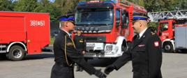 Strażak składa meldunek drugiemu strażakowi na tle samochodów strażackich.