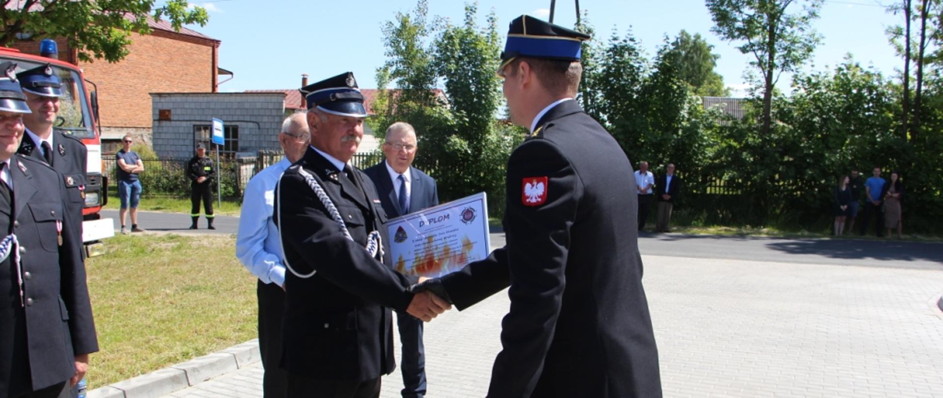 Zdjęcie wykonane przed strażnicą ochotniczej straży pożarnej w Goszczewicach. Na zdjęciu widać stojącego funkcjonariusza komendy miejskiej Państwowej Straży Pożarnej w Radomiu starszego kapitana Damiana Bechera. Funkcjonariusz w imieniu komendanta miejskiego dokonuje wręczenia prezesowi ochotniczej straży pożarnej w Goszczewicach pamiątkowego dyplomu z okazji uroczystości. 