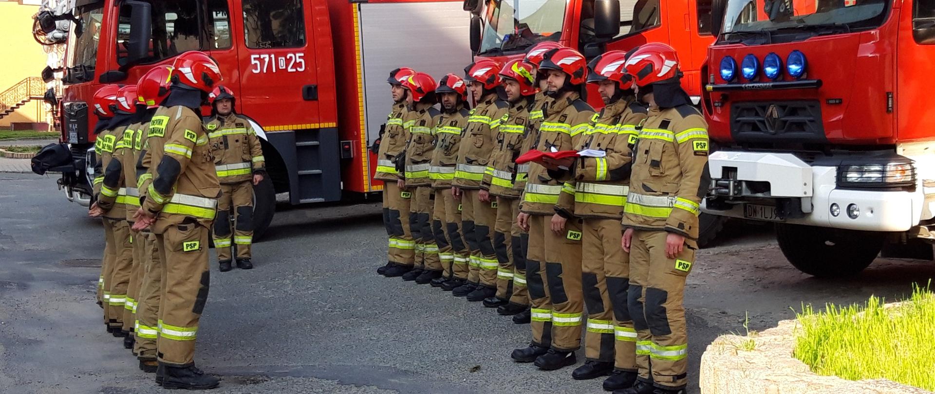 Strażacy w rzędzie przed samochodami bojowymi. Jeden ze strażaków trzyma na dłoniach złożoną flagę, przygotowaną do wciągnięcia na maszt. 