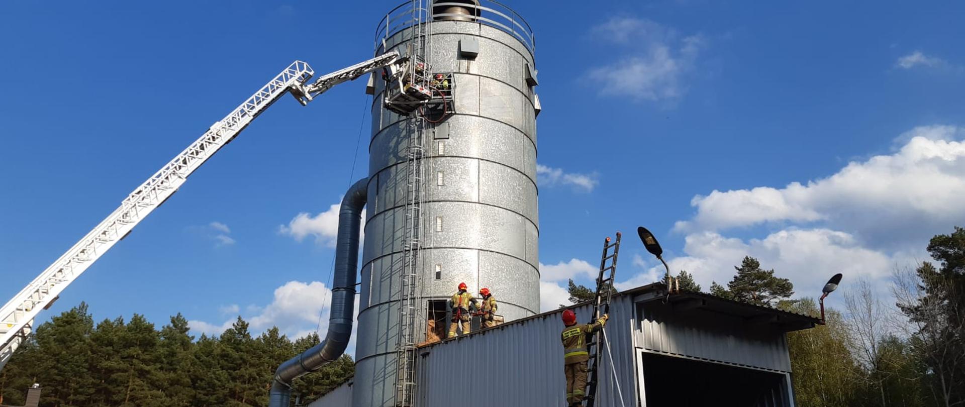 Strażacy gaszą pożar silos z podnośnika i wygarniają trociny.